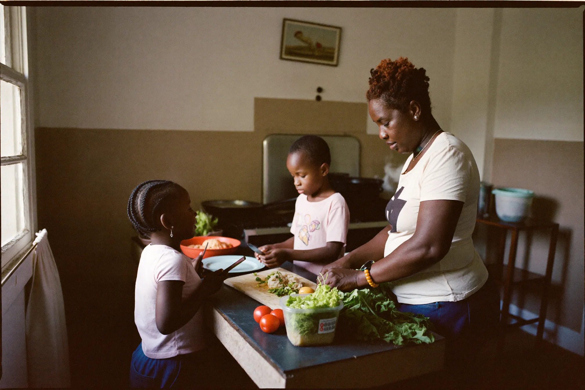 A mother and two children preparing a simple meal together in a modest South African kitchen with fresh vegetables on the counter